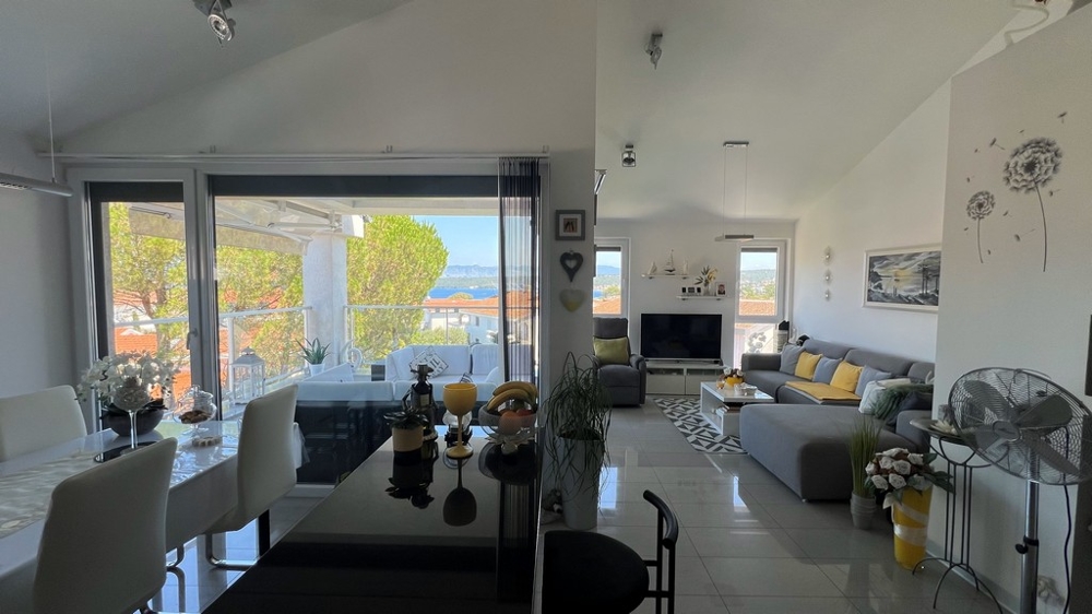Salon lumineux avec canapé gris, coin repas et grande façade vitrée donnant sur la terrasse avec vue mer.