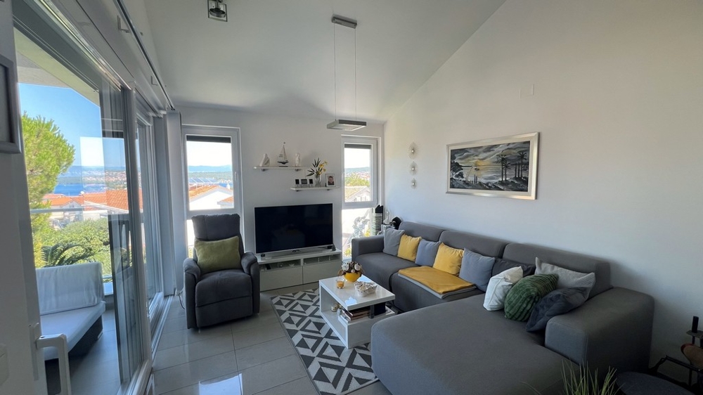 Salon moderne avec grande télévision, canapé gris et vue mer par de grandes fenêtres.