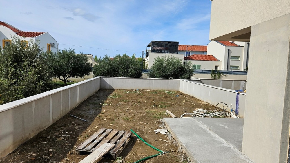 Espace extérieur au sol non stabilisé, mur bas et quelques arbres à l'arrière-plan.