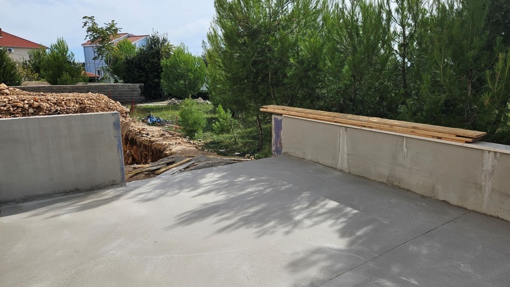 Espace extérieur en partie bétonné avec chantier et tranchée à côté de pins.