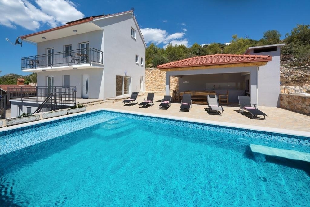 Vue sur la piscine à débordement, l'espace détente avec coin repas couvert et la maison.