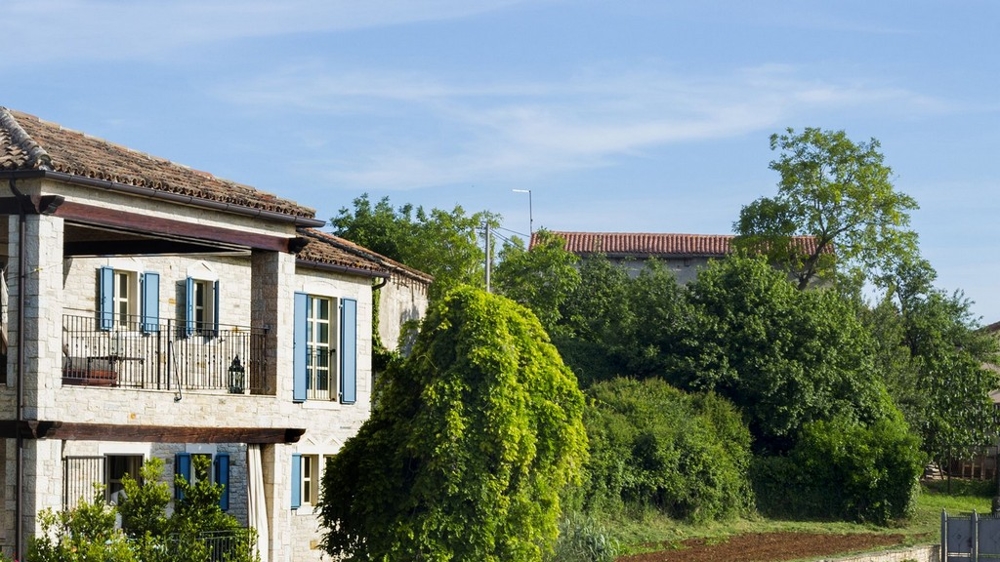 Maison en pierre avec volets bleus et balcon derrière des arbres