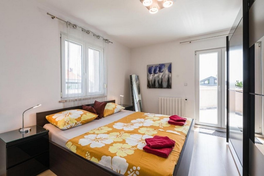 Chambre moderne dans un bien en Croatie, immeuble collectif avec vue mer, avec un lit double à la literie jaune, tables de chevet et œuvre d'art au mur, accès à une terrasse