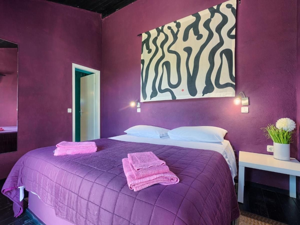 Une chambre dans un bien en Croatie, avec des murs violets, un grand tableau aux motifs abstraits, deux lampes de chevet et un linge de lit violet assorti