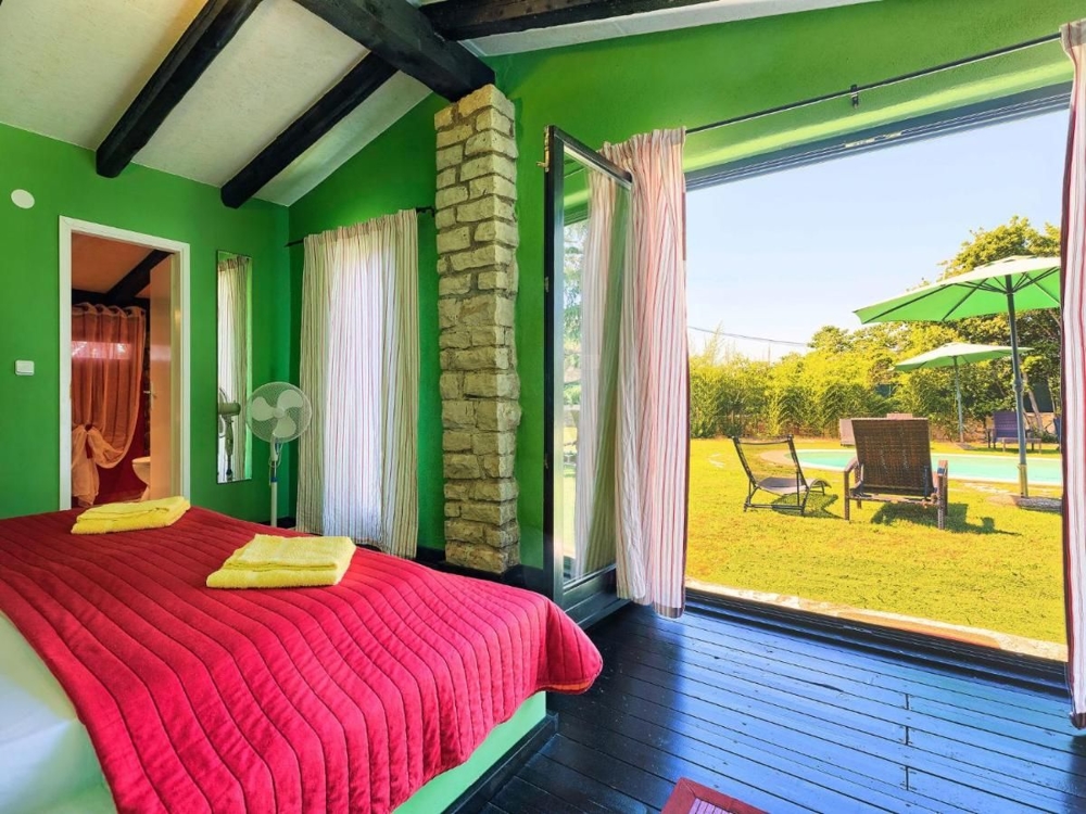 Chambre lumineuse avec vue sur le jardin, partie d'un bien en Croatie, avec des murs verts, un jeté de lit rouge et un accès à une terrasse avec des chaises longues et un parasol