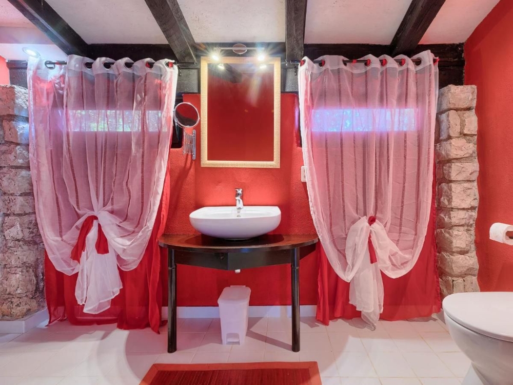 Salle de bains d'un bien en Croatie avec des murs rouges, des sanitaires blancs, un lavabo sur une table rustique et des rideaux rouges transparents aux fenêtres