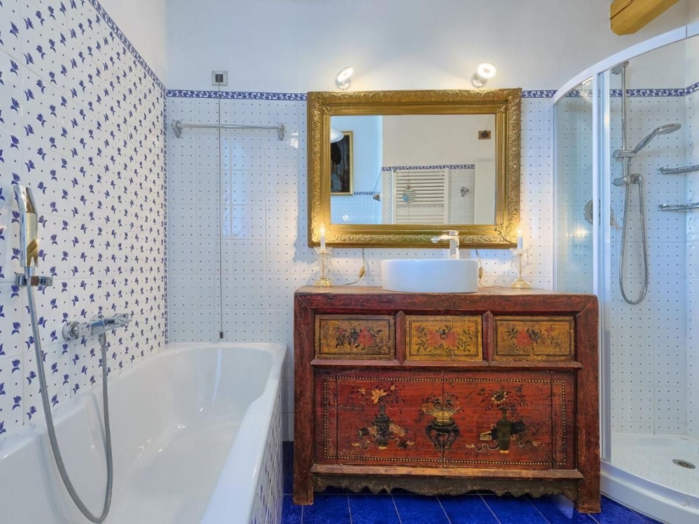 Une salle de bains dans un bien croate, ornée de carreaux bleus et blancs, d'une baignoire, d'un espace douche et d'un meuble ancien et coloré sous un miroir doré