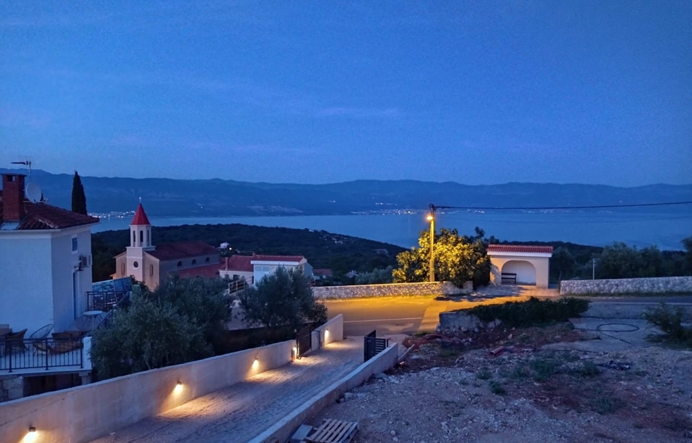 Vue depuis le bien en Croatie sur la mer de nuit, piscine au premier plan et village éclairé à l'arrière-plan.