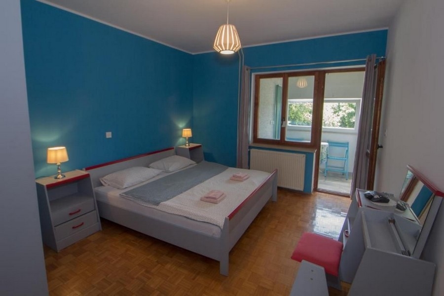 Une chambre aux murs peints en bleu, avec un grand lit double, deux tables de chevet avec lampes, une fenêtre et une porte donnant sur un balcon.