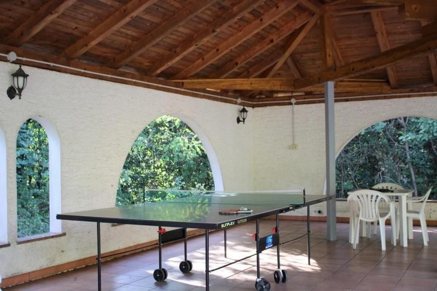 Un espace extérieur couvert avec une table de ping-pong au centre, des chaises en plastique blanc et un toit en bois. Les murs ont des ouvertures de fenêtres en arc donnant sur un environnement verdoyant.