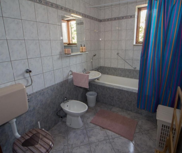 Une salle de bains avec carrelage blanc et gris, une baignoire, un lavabo, un bidet, un WC et un rideau de douche bleu. De petites fenêtres laissent entrer la lumière du jour.