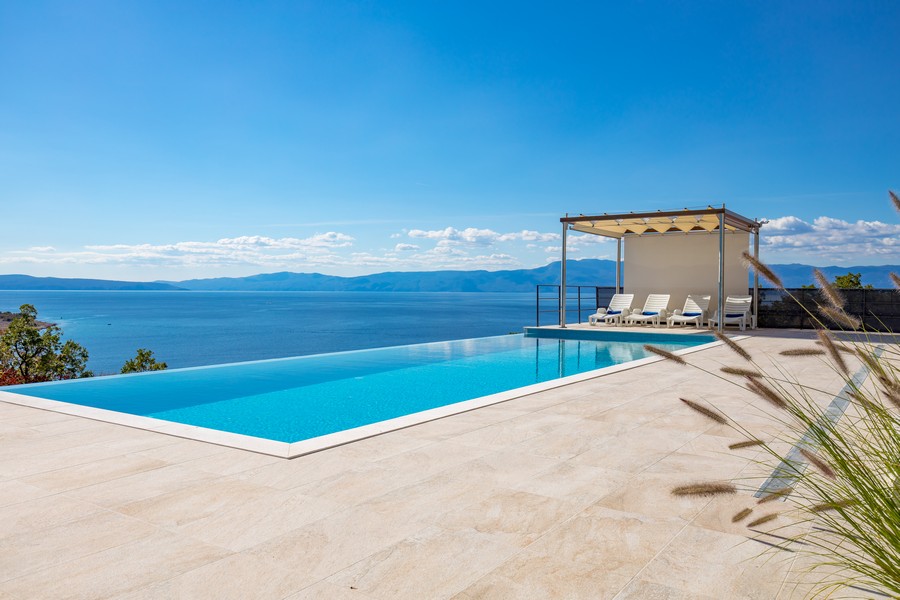 Piscine à débordement avec chaises longues, vue directe sur la mer et ciel bleu.