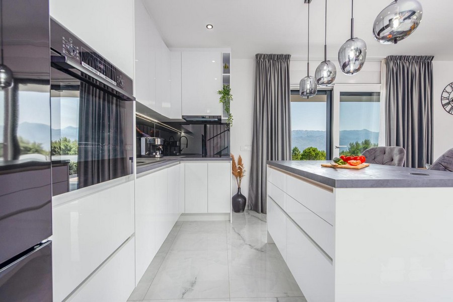 Cuisine blanche moderne avec îlot, suspensions et vue sur la nature par de grandes fenêtres.